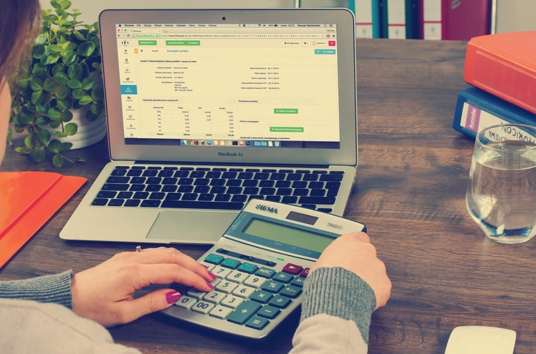 custos indiretos BDI: imagem mostra uma mulher em seu local de trabalho, sentada sem sua mesa, com o notebook aberto e usando uma calculadora