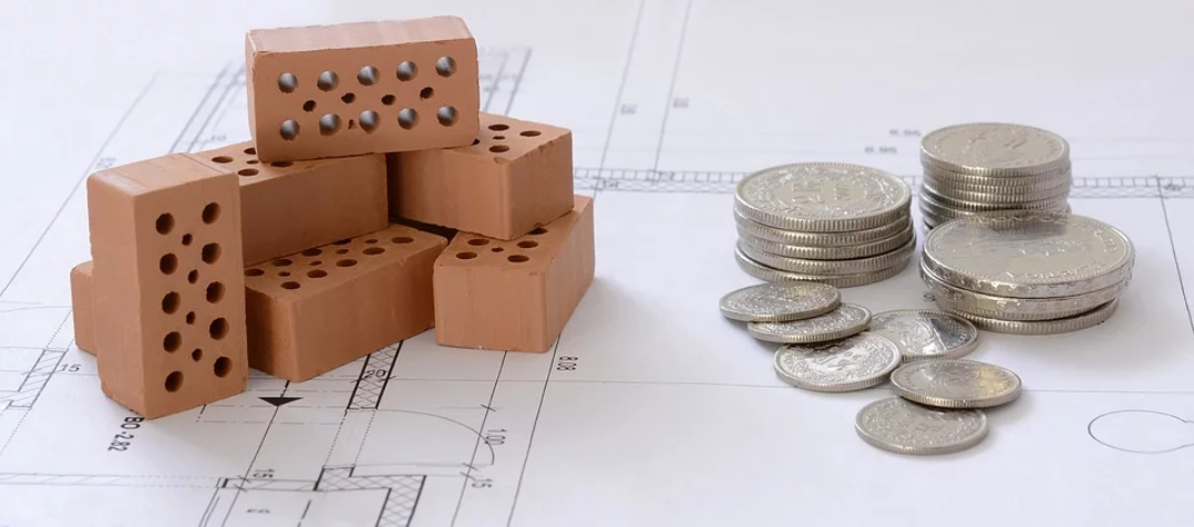 Crédito Imobiliário: ao fundo da imagem está o projeto gráfico de uma obra e em cima uns mini tijolos e do lado umas moedas empilhadas