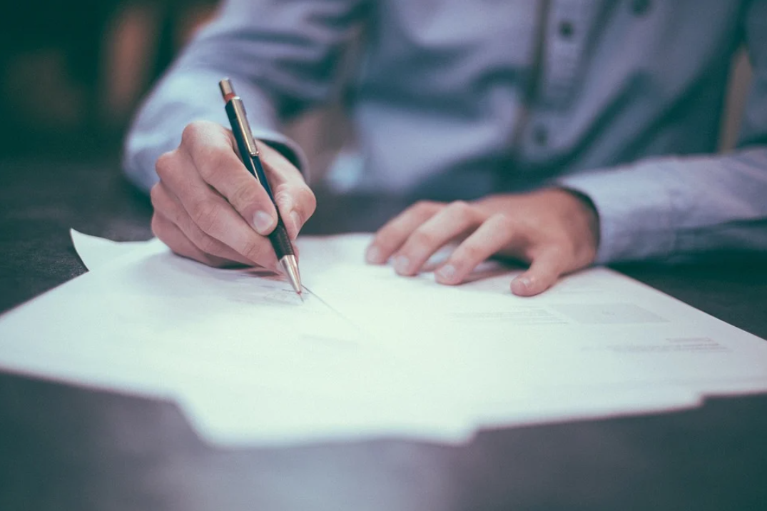 financiamento bancário: imagem mostra uma pessoa com as duas mãos sobre folhas de papel sobre uma mesa. A pessoa está segurando uma caneta e escrevendo.