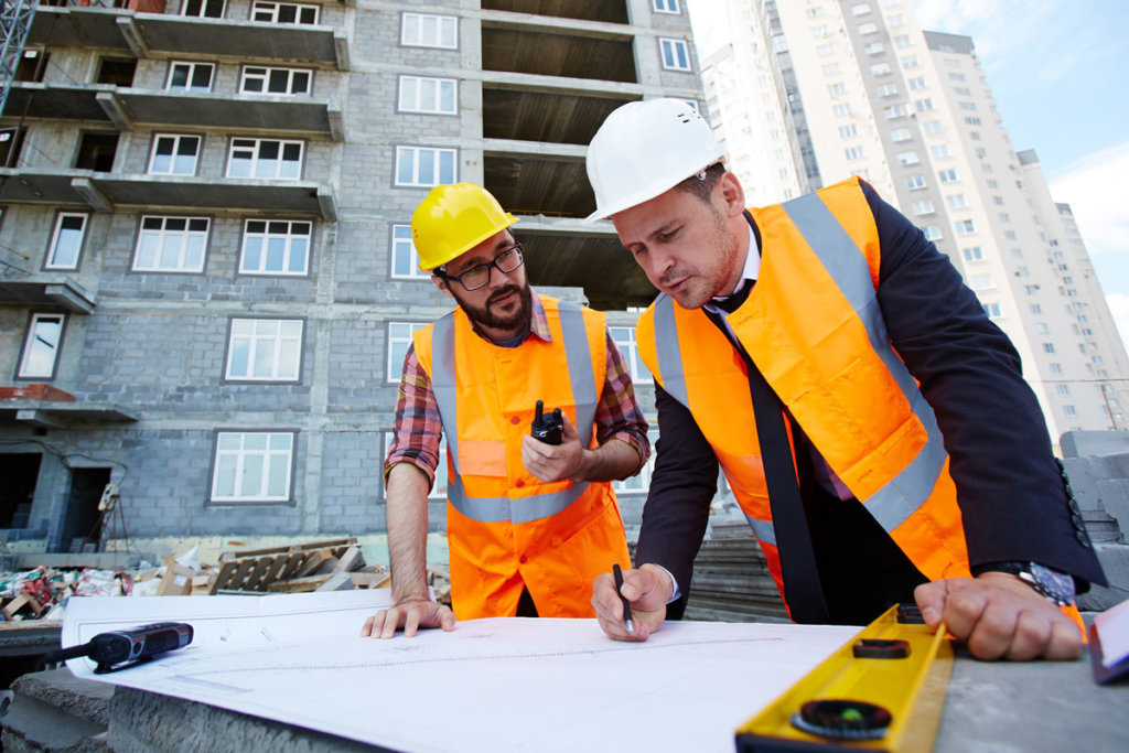 dois engenheiros analisando a planta de um prédio em frente a uma construção.